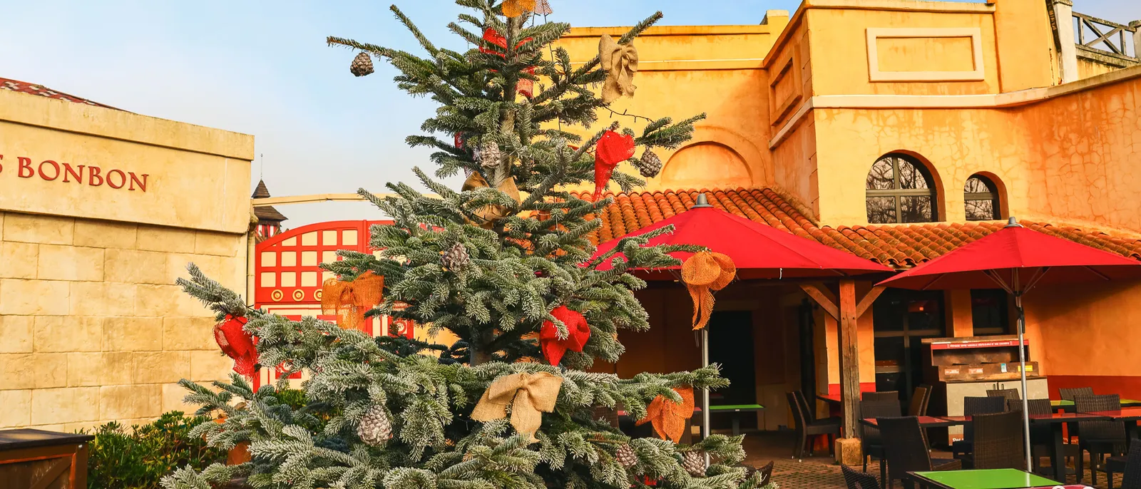 Sapin de Noël décoré de nœuds rouges et de rubans dorés devant des façades ocre et des parasols rouges