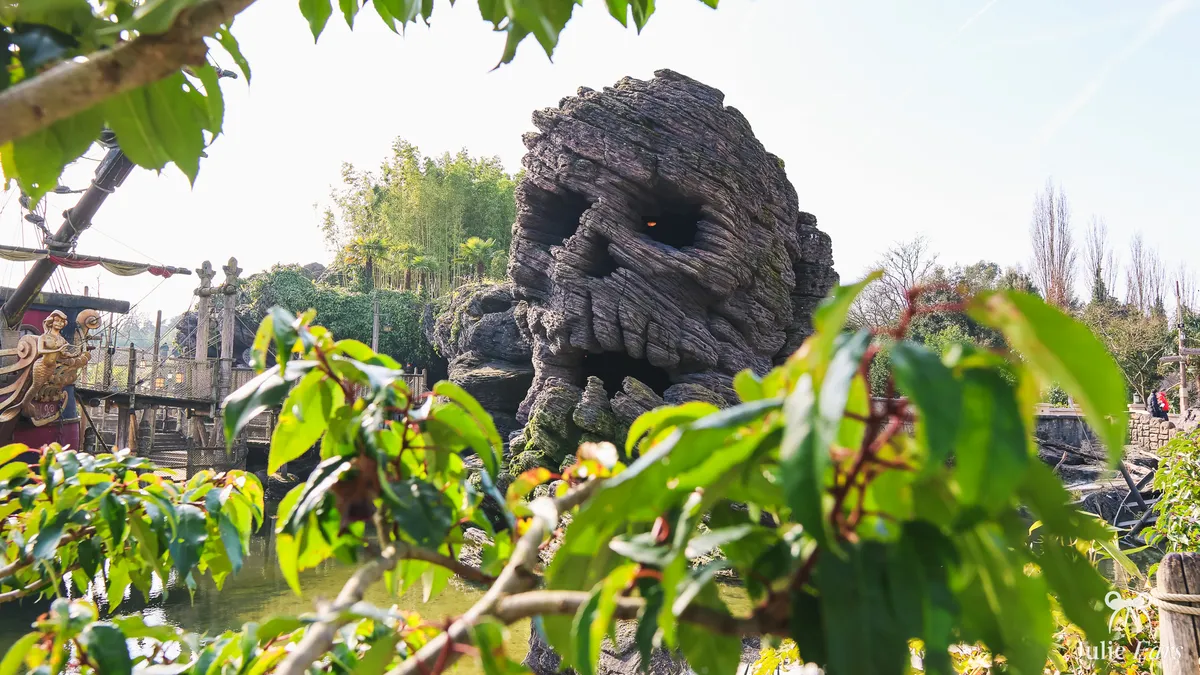 Skull Rock, rocher sculpté en forme de crâne sur Adventure Isle à Disneyland Paris, vu à travers un feuillage vert, avec la proue d’un galion de pirates et sa figure de proue en sirène sur la gauche.