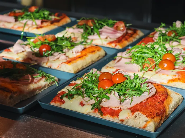 Parts de pizza rectangulaires garnies de jambon, roquette et tomates cerises, sur des plateaux au Stark Factory.