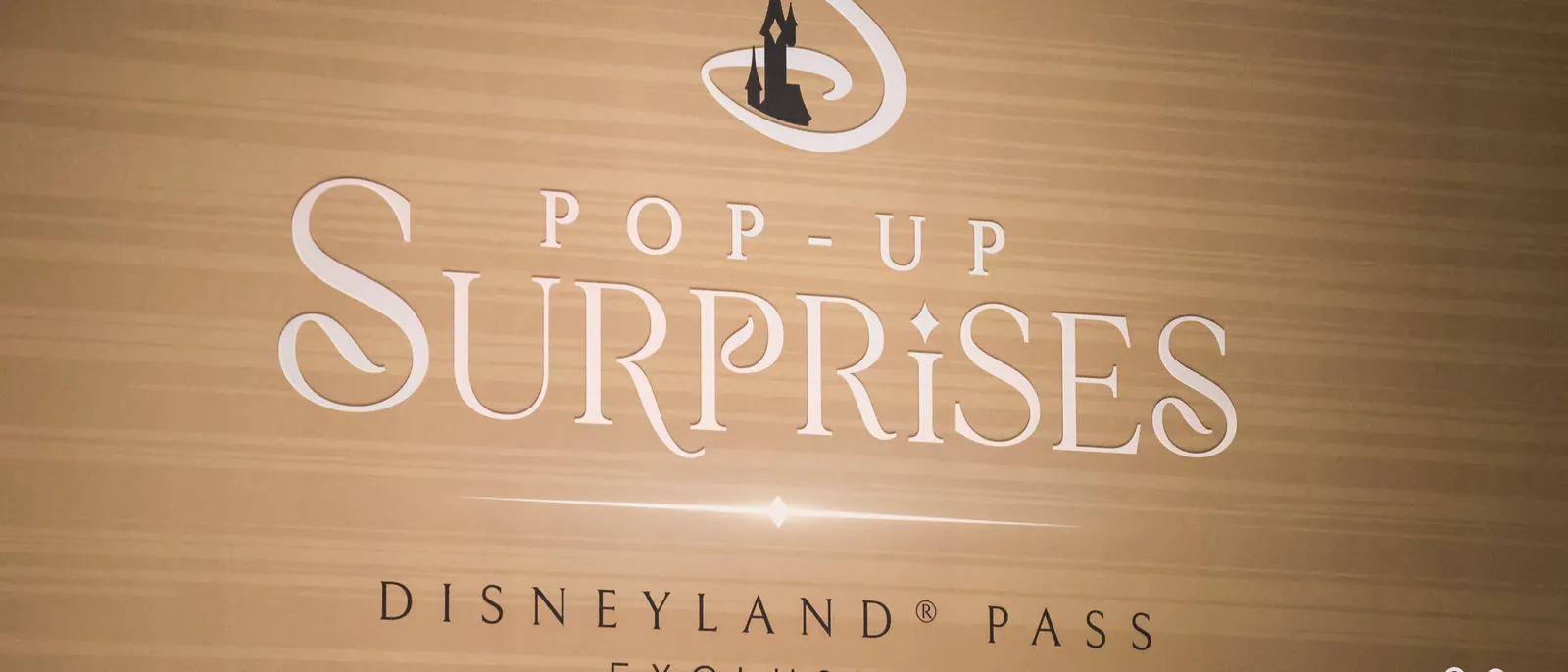 Enseigne « Pop-Up Surprises Disneyland Pass Exclusive » avec le logo château, sur un mur en bois clair