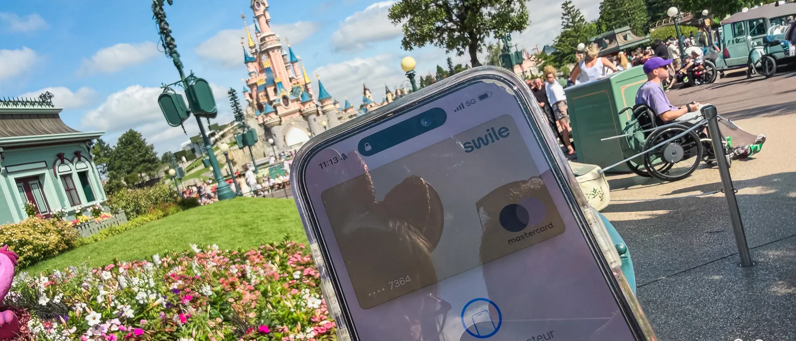 Smartphone affichant l’application Swile prête à payer, devant le château de la Belle au Bois Dormant à Disneyland Paris.
