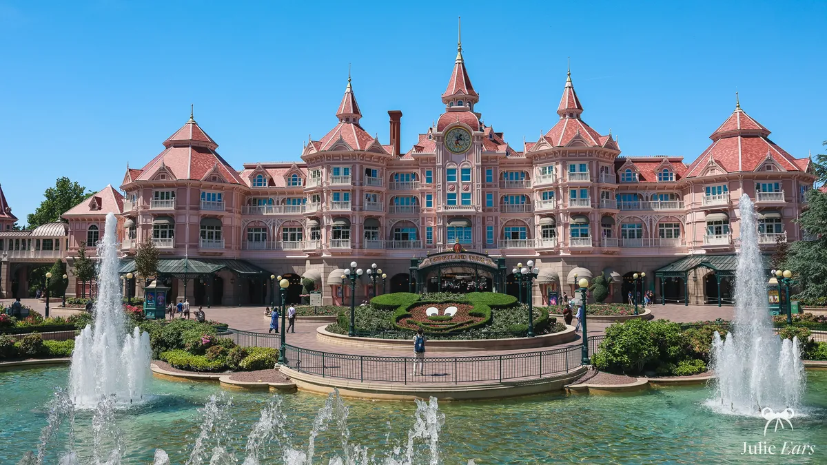 Disneyland Park entrance beneath the pink Disneyland Hotel facade at Disneyland Paris, with fountains and a Mickey-shaped flower bed