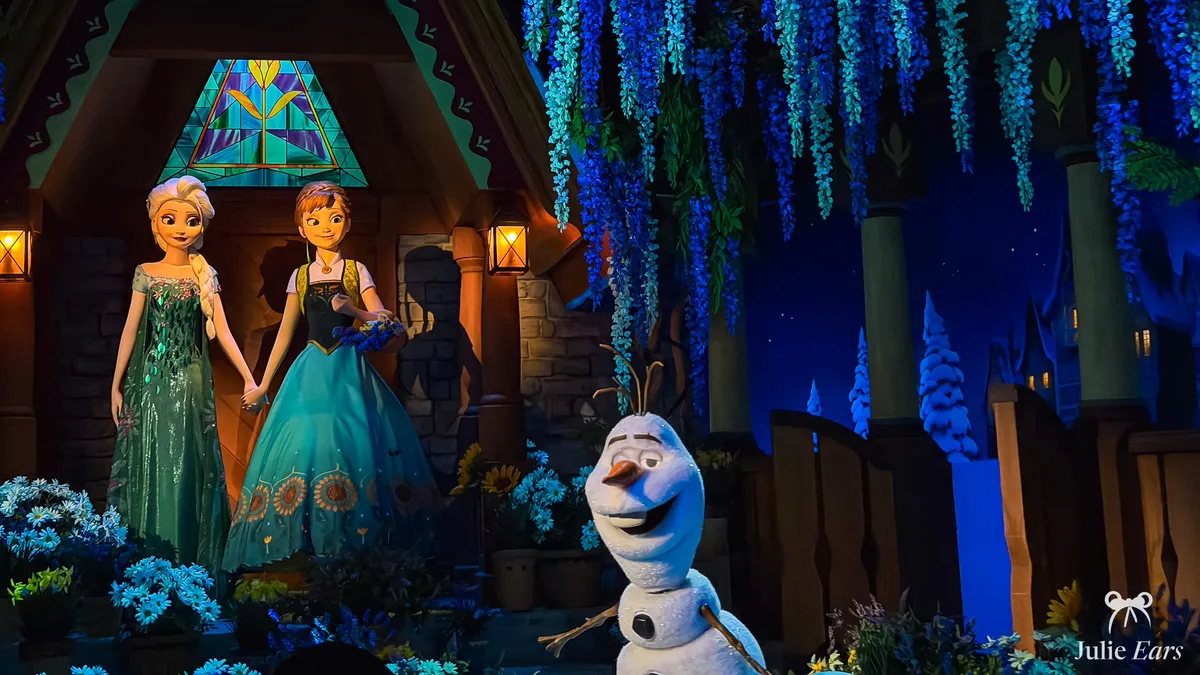 Final scene of the Frozen Ever After ride at Disney Adventure World: Elsa and Anna hand in hand in front of an Arendelle house, with Olaf in the foreground and blue wisteria