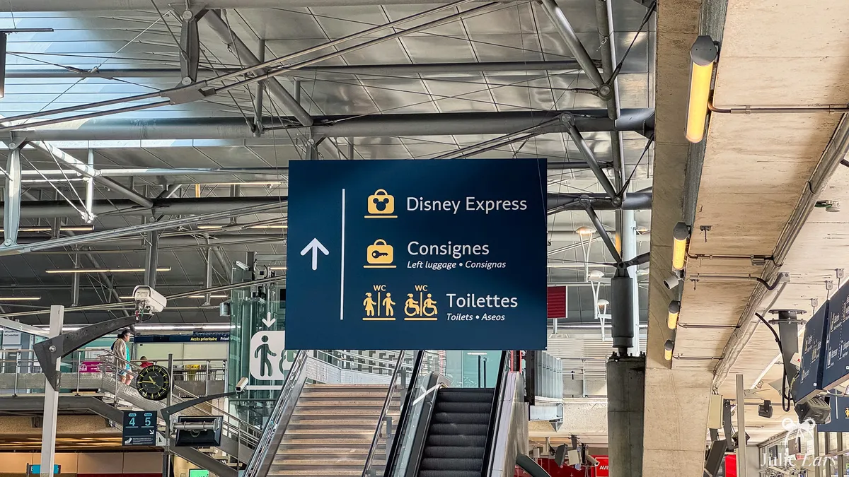 Panneau de direction bleu dans la gare de Disneyland Paris indiquant le Disney Express, les consignes à bagages et les toilettes, au-dessus des escalators.