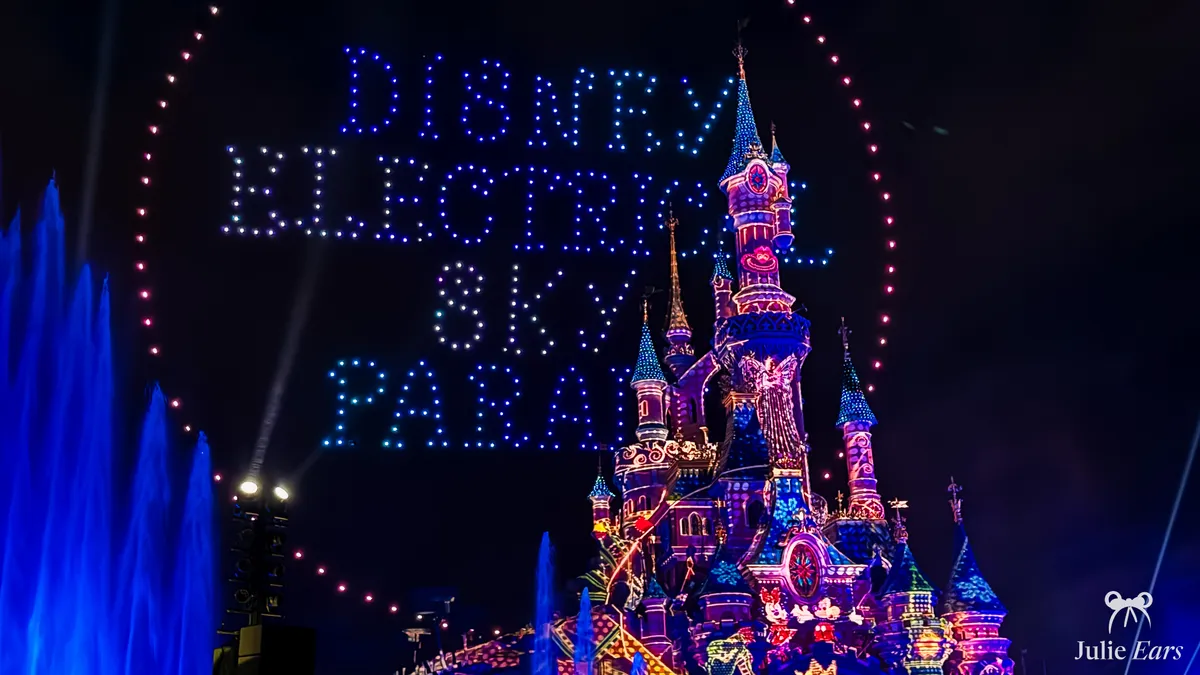 Drones spelling "Disney Electrical Sky Parade" in a circle of light above Sleeping Beauty Castle at Disneyland Park, with blue fountains in front