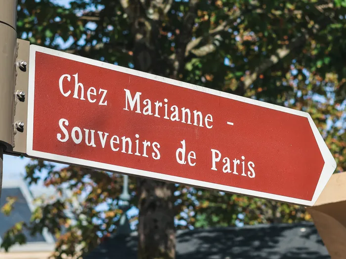 Red arrow-shaped signpost pointing to "Chez Marianne, Souvenirs de Paris", fixed to a post in front of the trees, at Disneyland Paris.
