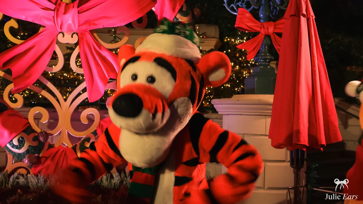 Tigger in a Santa hat and striped scarf at night, by a golden gate with a big pink bow and fairy lights, at Christmas Night 2023 at Disneyland Paris