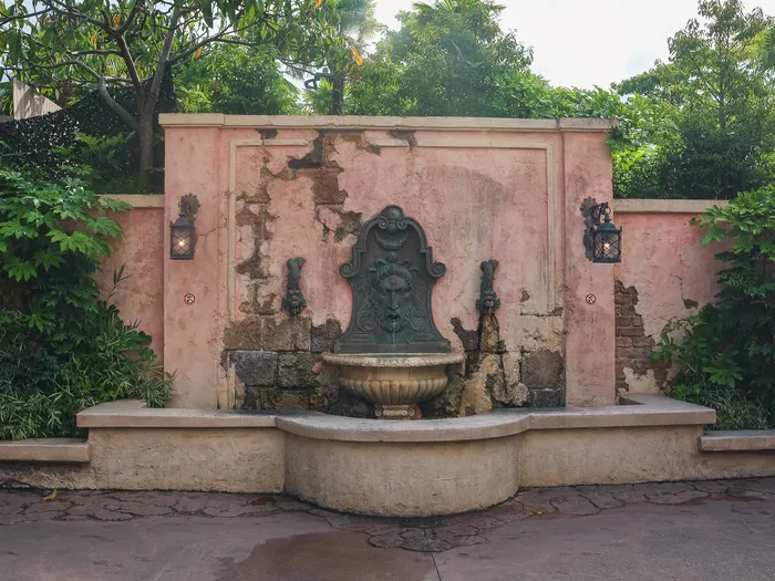 Fontaine murale en pierre sur un mur rose patiné, entourée de verdure dans le patio de Captain Jack’s à Adventureland.