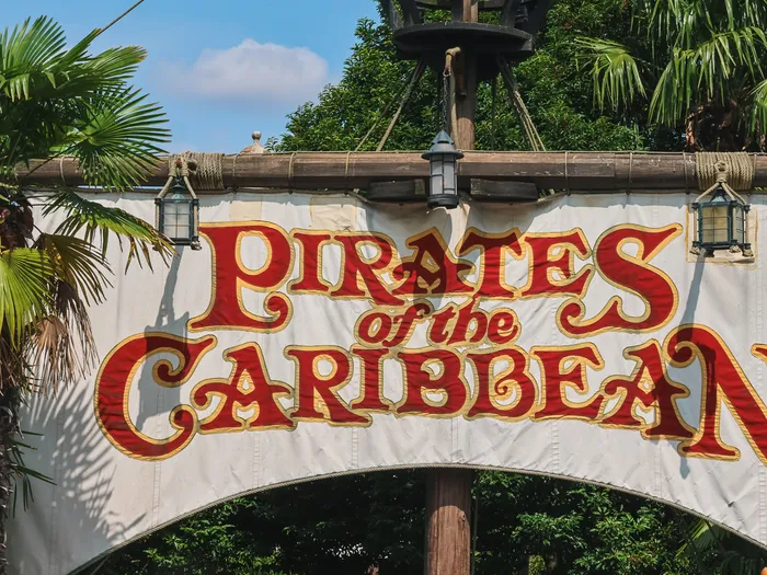Bannière blanche aux lettres rouges « Pirates of the Caribbean » à l’entrée de l’attraction, entourée de palmiers.