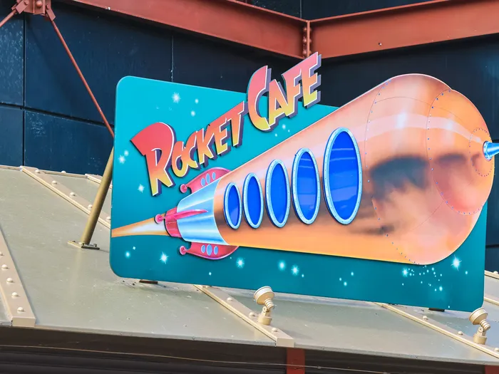 Rocket Café sign in Discoveryland, Disneyland Paris: a copper rocket with blue portholes crossing a turquoise starry sky, the name in red