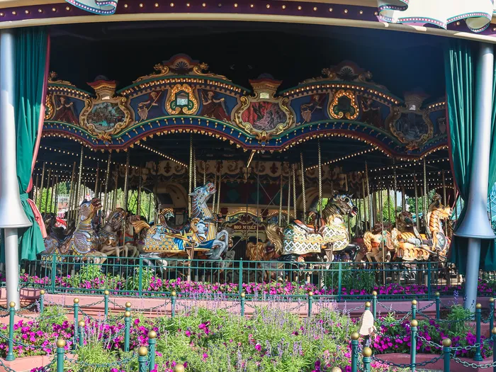 Gros plan sur un cheval de bois blanc harnaché du Carrousel de Lancelot, fleurs roses derrière la barrière verte.
