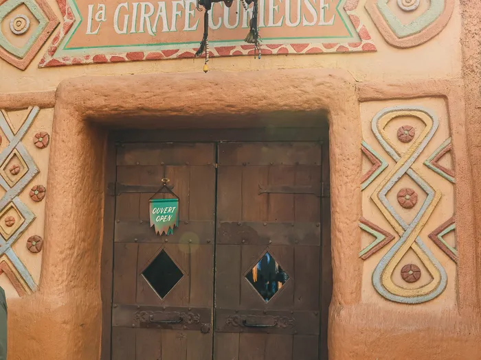 Colourful patterned daub facade of the La Girafe Curieuse shop at Disneyland Paris, with its sign above wooden doors and an "Ouvert / Open" sign