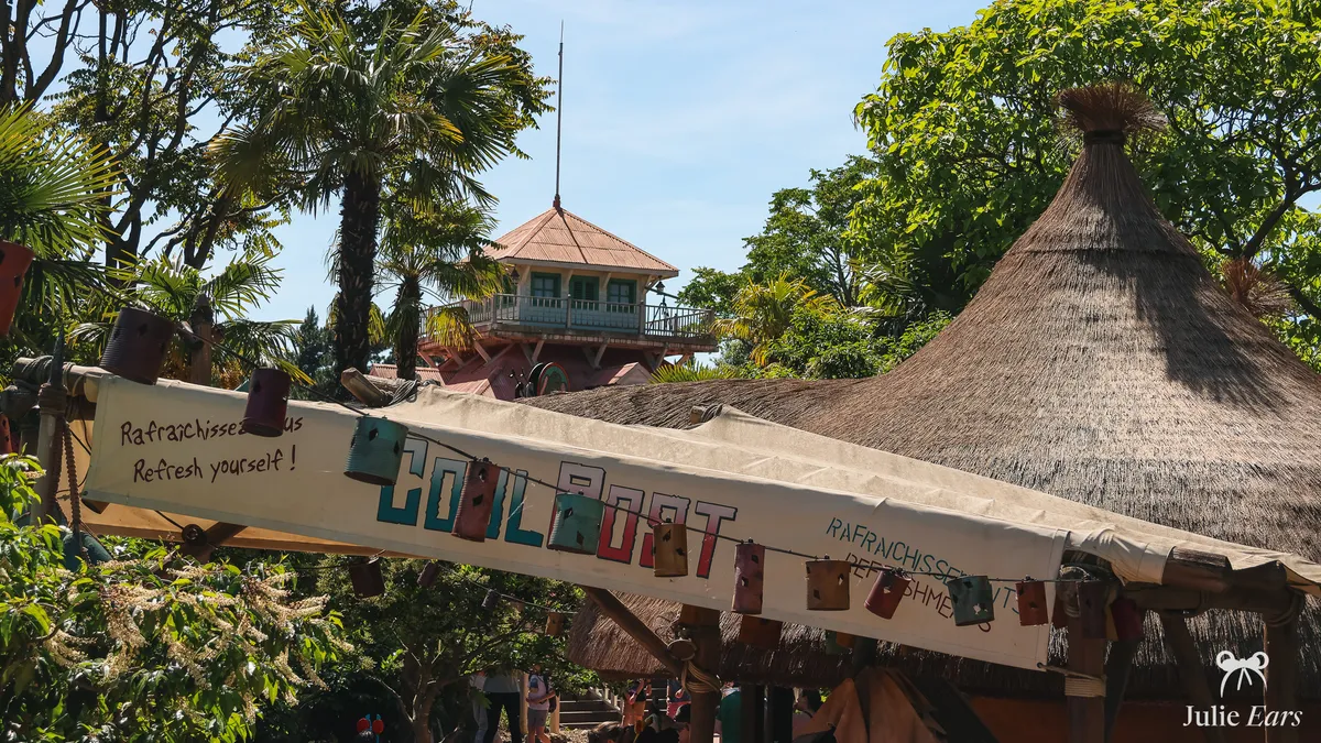 Cool Post kiosk in Adventureland, Disneyland Park, with its banner inviting you to cool off, a tin can garland and a thatched roof under palms