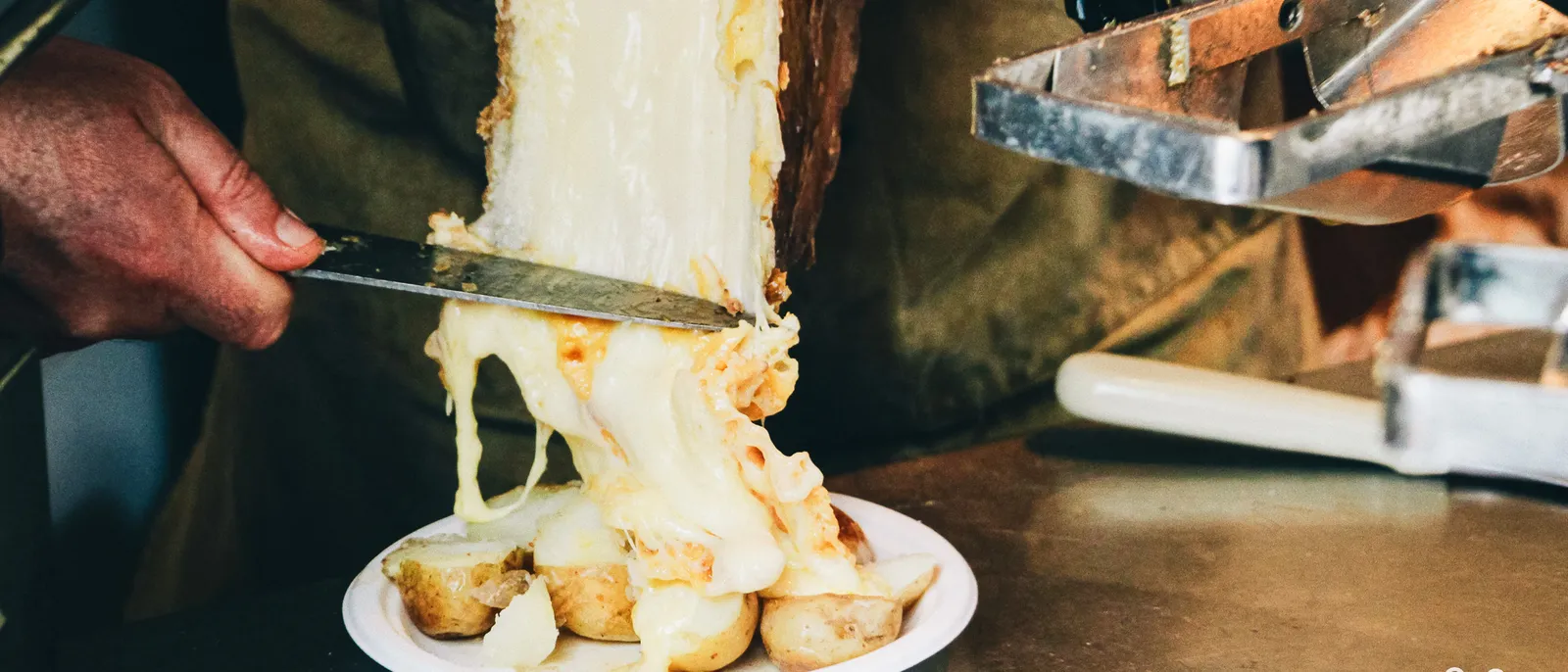 Demi-meule de raclette fondue raclée au couteau sur des pommes de terre, filet de fromage coulant dans l’assiette