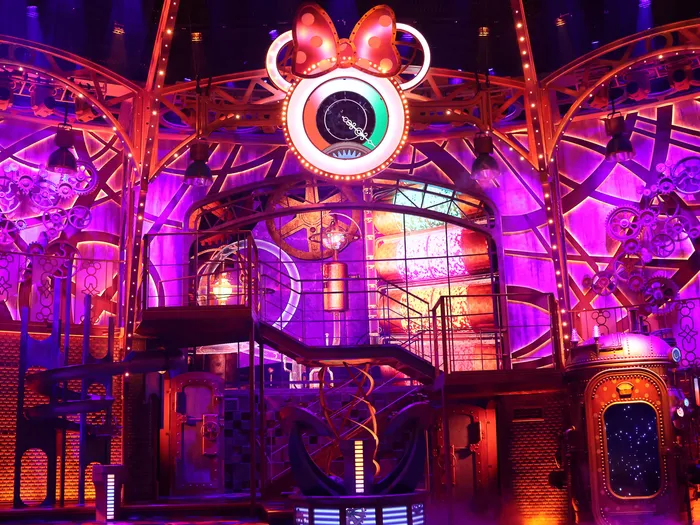 Scène industrielle éclairée de violet avec grande roue et nœud de Minnie lumineux, décor du spectacle Fabrique des Rêves au Parc Disney Adventure World.
