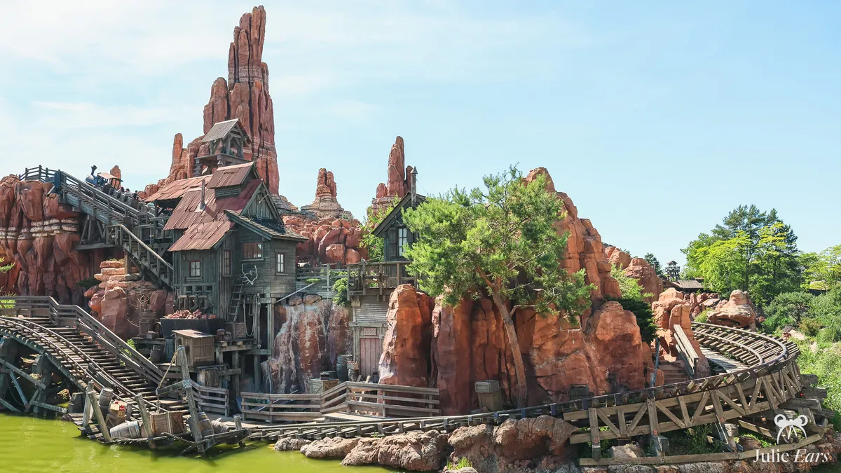 Big Thunder Mountain at Disneyland Paris: red rock peaks, wooden mining town houses and a train on the lift hill by the lake, under a blue sky