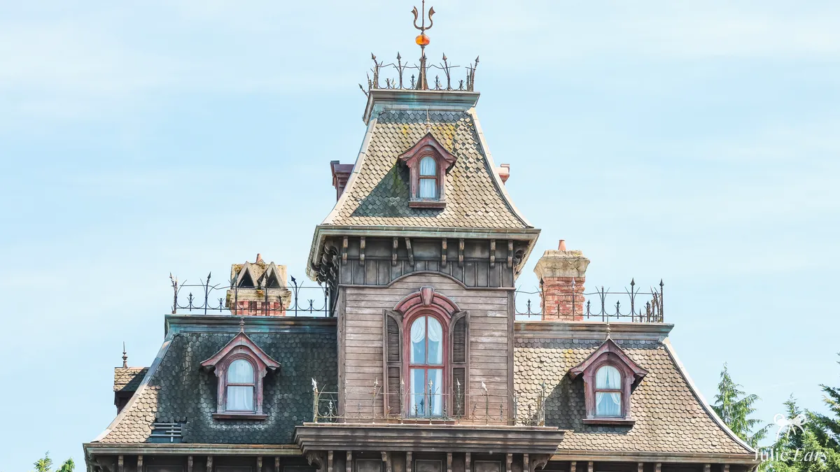 Top of Phantom Manor in Frontierland, Disneyland Paris: scalloped roof, dormer windows, brick chimneys and a wrought-iron finial against a blue sky