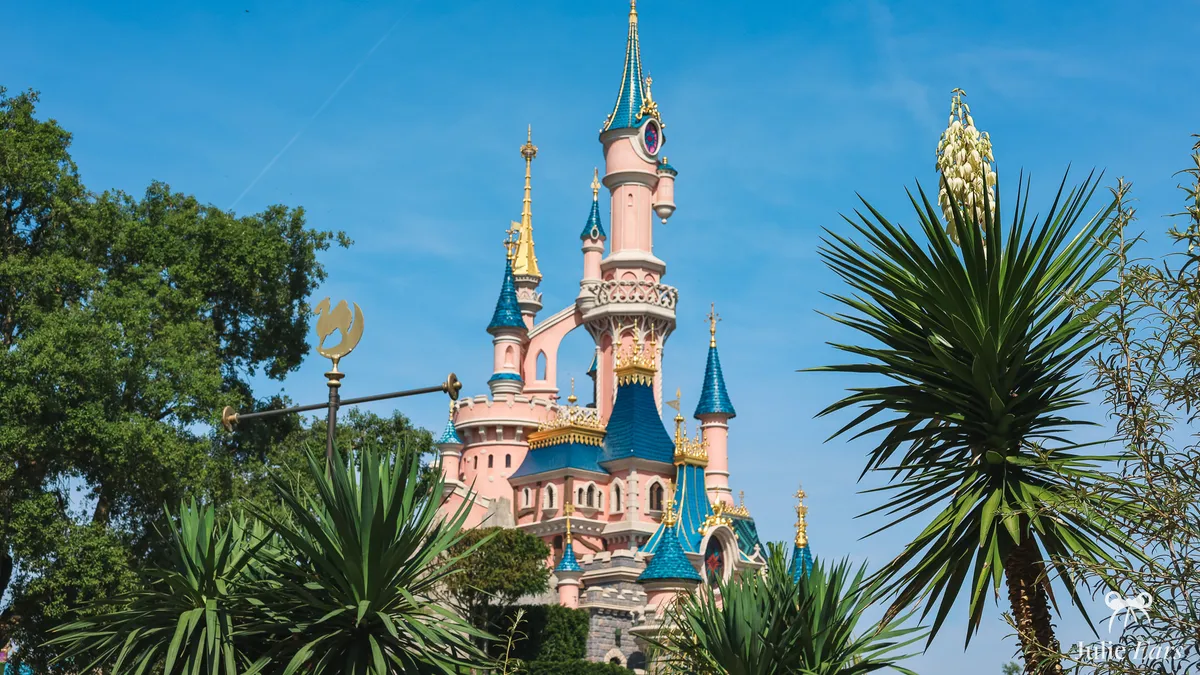 Sleeping Beauty Castle at Disneyland Paris seen from the Adventureland side, between yuccas and trees, with a golden camel post in front