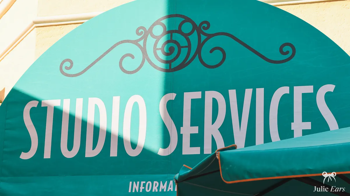 Turquoise arched awning of Studio Services at Disney Adventure World, crossed by a diagonal shadow, with white lettering beneath a black wrought-iron style pattern and a parasol in the foreground