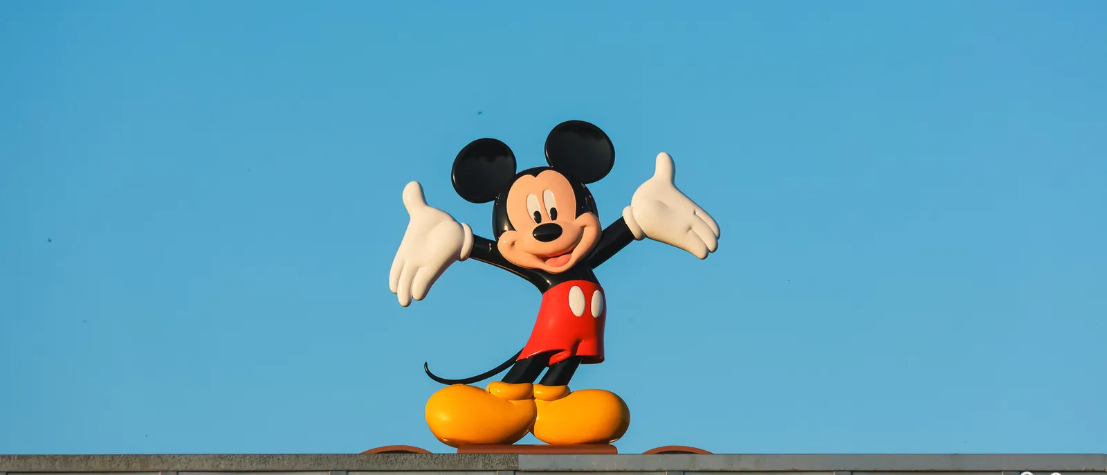 Statue de Mickey aux bras grands ouverts perchée sur un toit, se détachant sur un ciel bleu au Disney Village