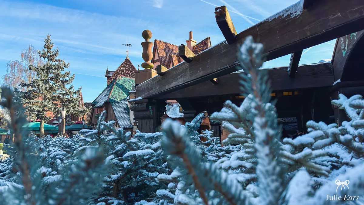 Snow-covered young fir trees in front of a snowy timber frame and the pointed Fantasyland roofs, under a blue winter sky at Disneyland Park