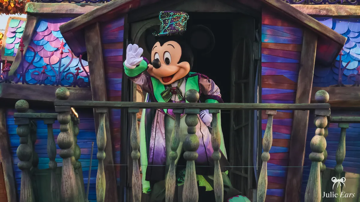 Mickey Mouse en costume d’Halloween, redingote violette et haut-de-forme à paillettes, salue depuis une maisonnette en bois colorée, pendant la saison d’Halloween à Disneyland Paris.