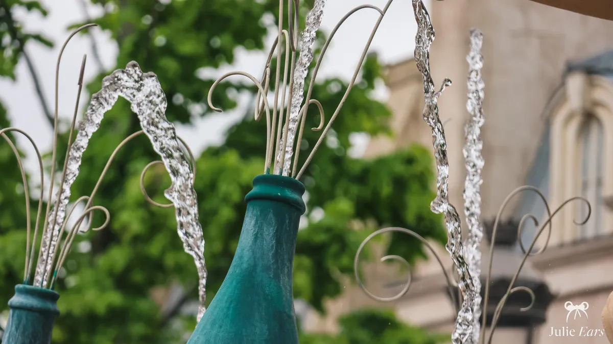 Water jets shooting from green champagne bottles between wrought-iron scrolls, on a fountain in La Place de Rémy at Disney Adventure World