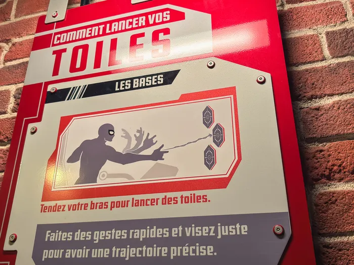Panneau explicatif « Comment lancer vos toiles » sur mur de brique dans la file d’attente de l’attraction Spider-Man W.E.B. Adventure.