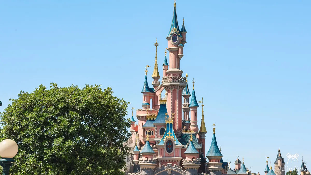 Sleeping Beauty Castle at Disneyland Paris, pink towers and blue and gold roofs under a clear blue sky, with a large tree and a globe lamppost