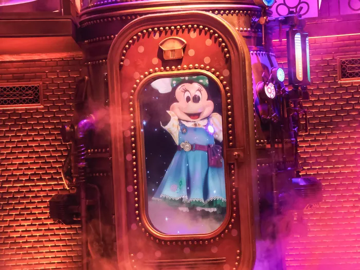 Minnie en robe bleue apparaît derrière un hublot de machine à vapeur dorée sur la scène du spectacle Minnie's Dream Factory