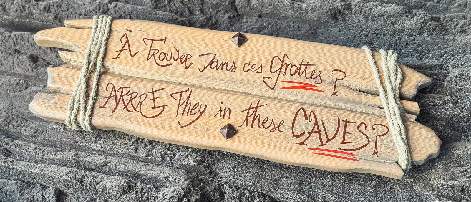 Pancarte en bois gravée "À trouver dans ces grottes ?" posée sur un rocher, indice de chasse au trésor.