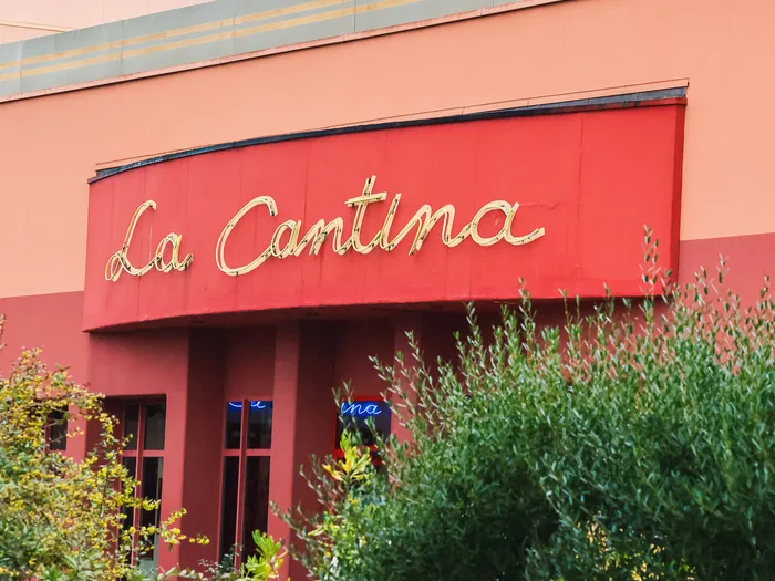Enseigne néon dorée La Cantina sur la façade rouge du restaurant, entourée de végétation et d’un rocher.