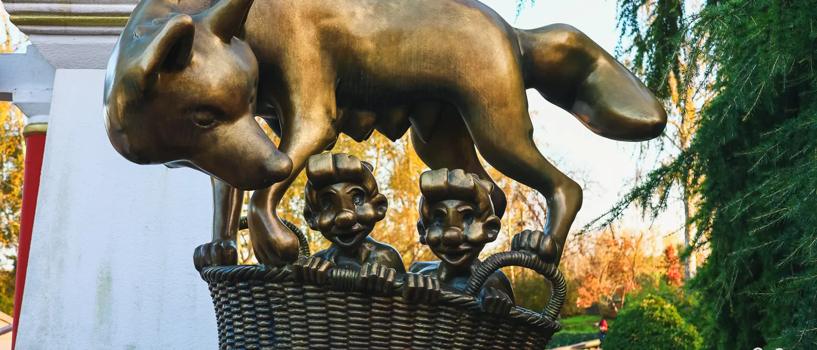 Statue en bronze d'une louve surplombant un panier en osier où se tiennent deux enfants, arbres roux à l'arrière-plan