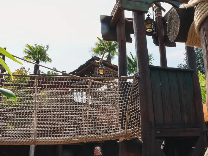 Pont de filet suspendu et tour de bois de l’aire de jeux Plage des Pirates, entouré de palmiers sous le soleil