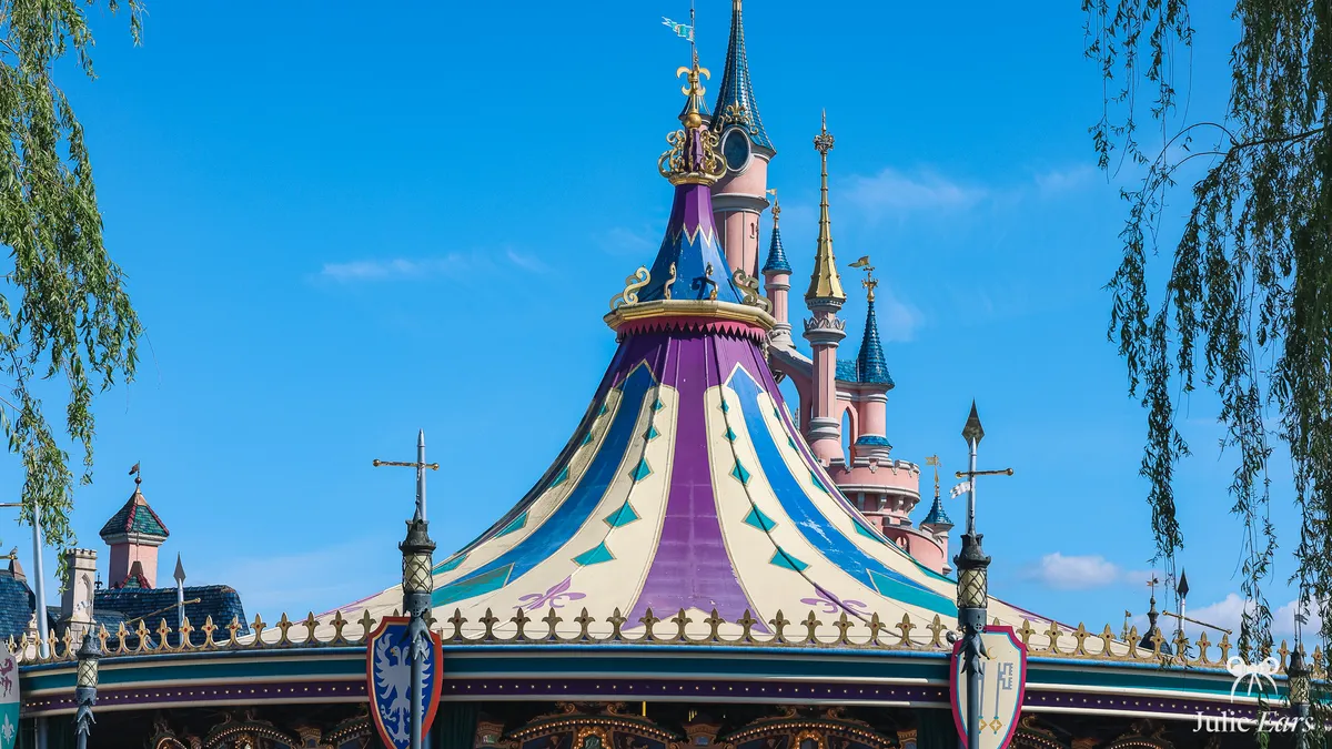 Purple, blue and cream canopy of Le Carrousel de Lancelot, with Sleeping Beauty Castle just behind, under a blue sky, in Fantasyland
