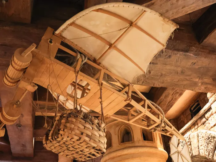 Wooden flying machine with a sail and wicker basket hanging from the ceiling beams, part of the Merlin l’Enchanteur shop decor at Disneyland Paris.