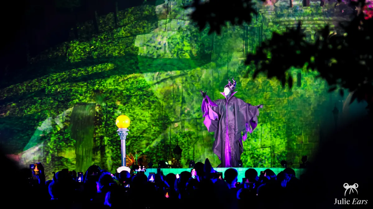 Maleficent, arms outstretched on stage in front of Sleeping Beauty Castle covered in green foliage projections, at Villainous Night 2023