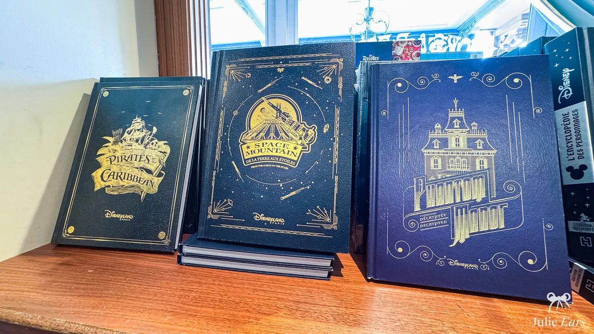 Three Disneyland Paris coffee table books on a shelf, in the colours of the Pirates of the Caribbean, Space Mountain and Phantom Manor rides.