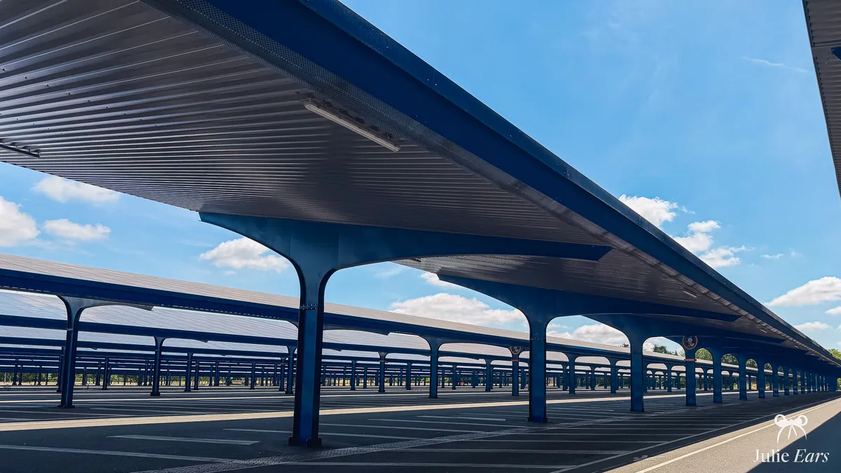 Rows of blue solar-panel canopies on arched pillars in the empty Disneyland Paris visitor car park, under a blue sky dotted with clouds