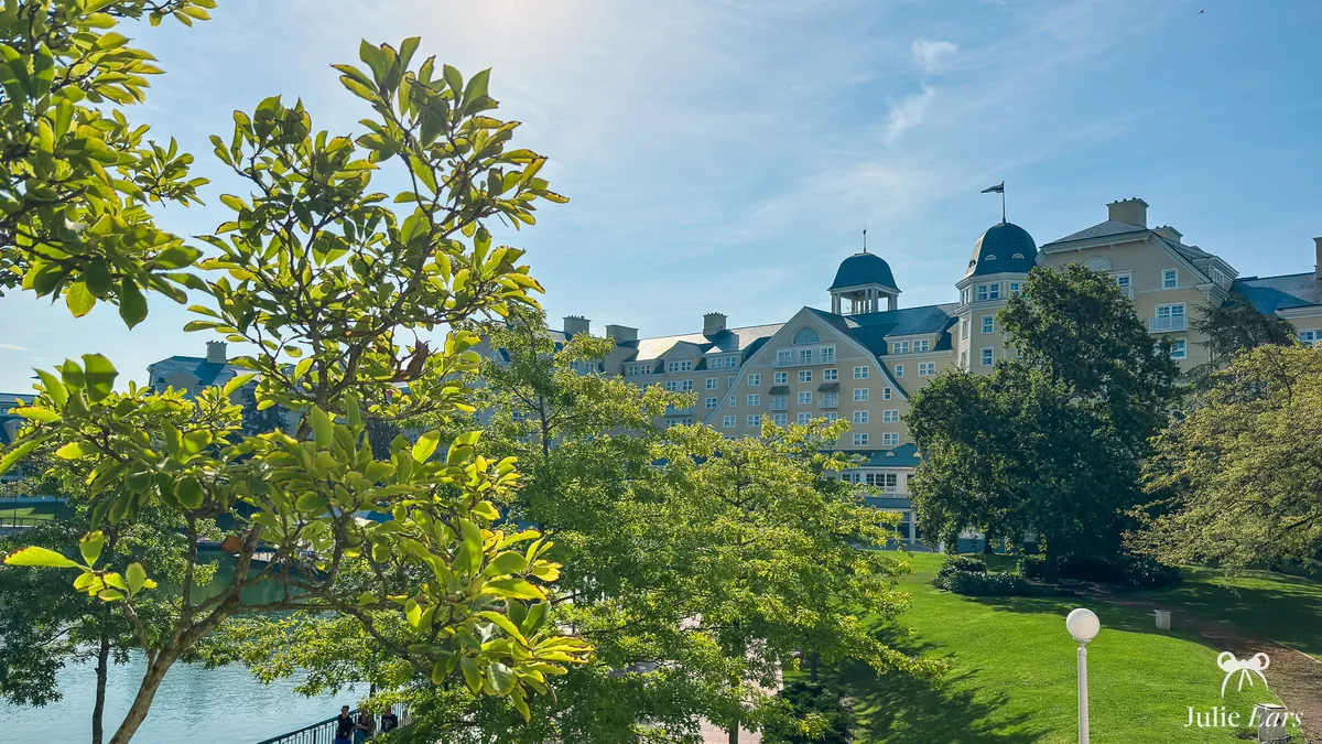 Disney Newport Bay Club on the shore of Lake Disney, seen through foliage: pale yellow facade, grey-blue roofs and domes, sunny lawn