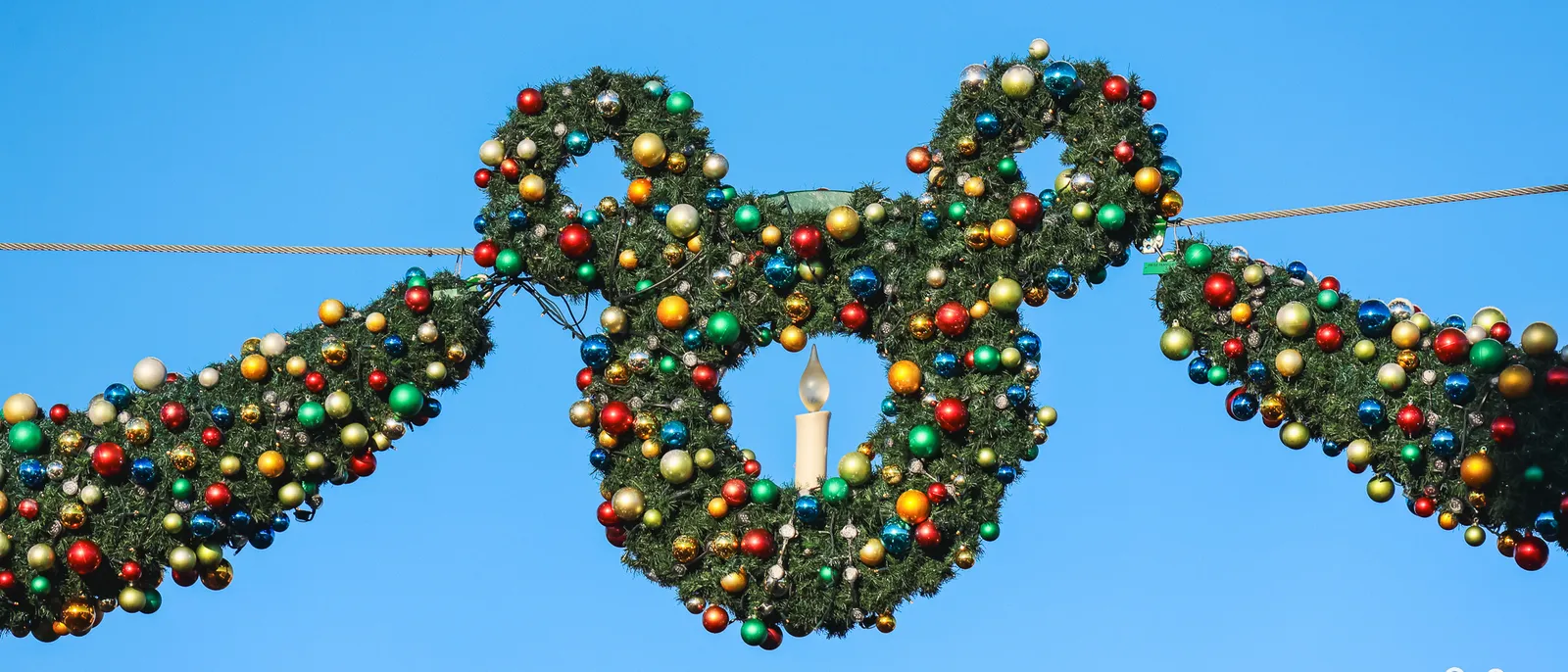 Grande couronne de sapin en forme de tête de Mickey, couverte de boules multicolores, suspendue sur ciel bleu.