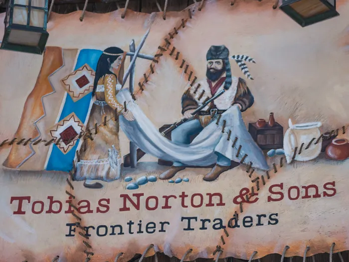 Enseigne en cuir peint « Tobias Norton & Sons Frontier Traders » figurant un trappeur et une trappeuse à Frontierland