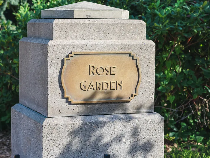 Grey stone post with a gold Art Deco plaque engraved "Rose Garden", in front of green shrubs, in a garden at Disneyland Paris.