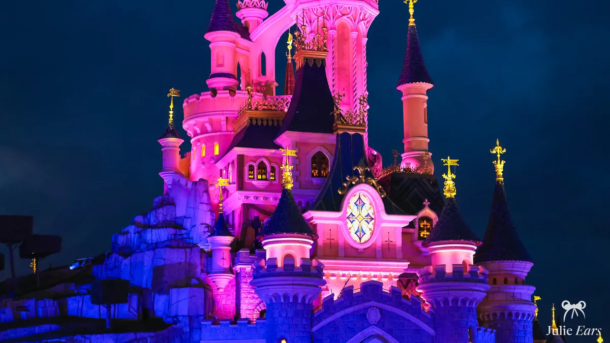 Sleeping Beauty Castle lit in pink and purple at nightfall, rose window glowing and gold weathervanes against a midnight blue sky, Disneyland Park