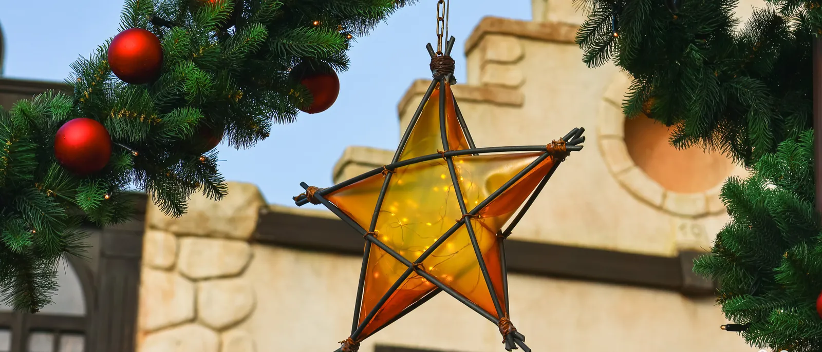 Étoile lumineuse orange suspendue entre deux guirlandes de sapin et boules rouges, devant une façade de pierre claire