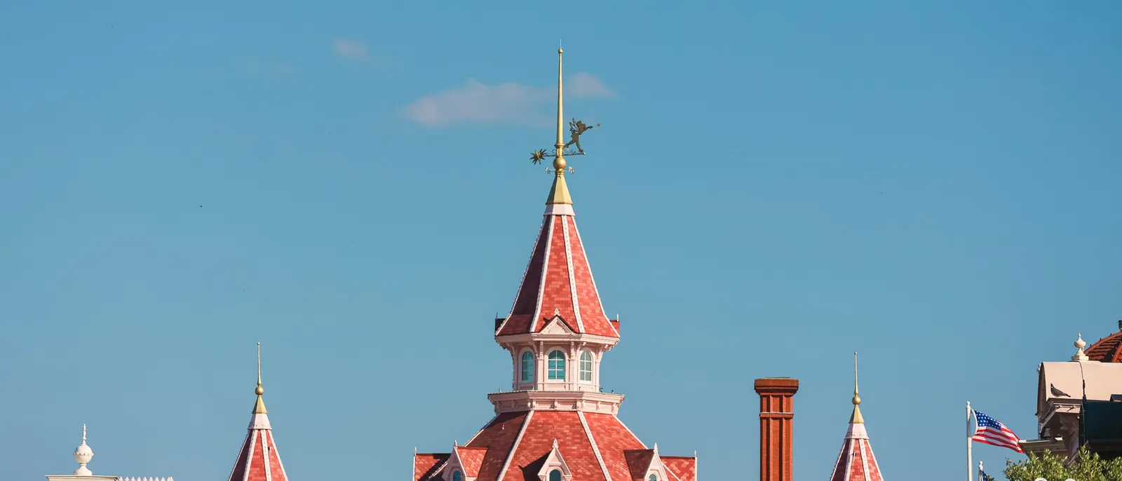 Les toits rouges à tourelles du Disneyland Hotel avec girouette dorée et drapeau américain, sous un ciel bleu.