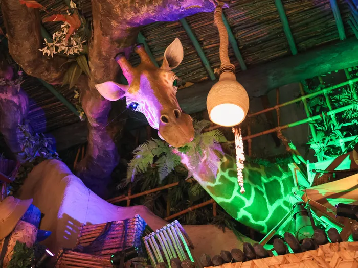 The decorative giraffe of La Girafe Curieuse reaching for a gourd lamp among rocks and plants lit in green and purple, Disneyland Paris