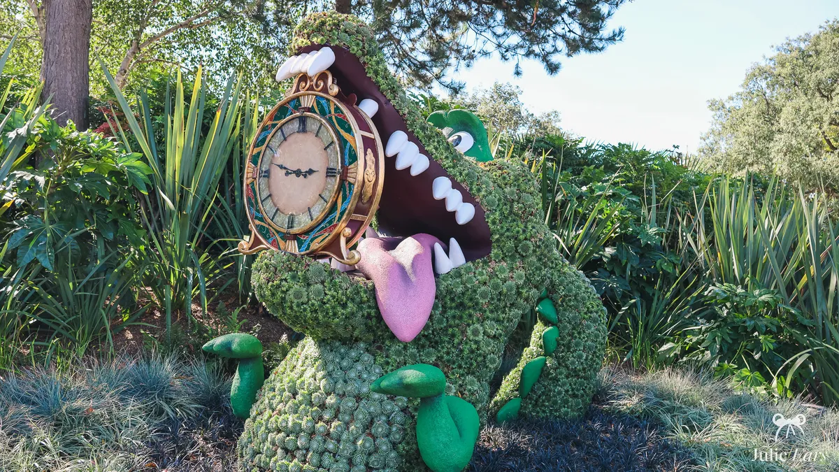 Topiary of Tick-Tock, the crocodile from Peter Pan, jaws open and pink tongue out, a clock in its teeth, in a flower bed at Disneyland Park