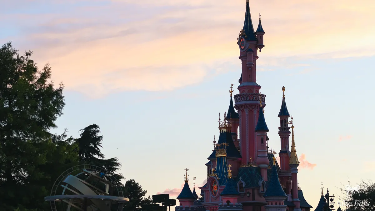 Pink spires of Sleeping Beauty Castle at sunset under a pastel peach and blue sky, with the Discoveryland astrolabe and tall dark trees on the left