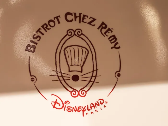 Close-up of the "Bistrot Chez Rémy" logo printed in brown on beige crockery, with a chef’s hat, rat whiskers and the Disneyland Paris signature.
